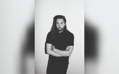 Rasmus Syman, ein Mann mit langen Haaren und Bart, steht mit verschränkten Armen vor hellem Hintergrund