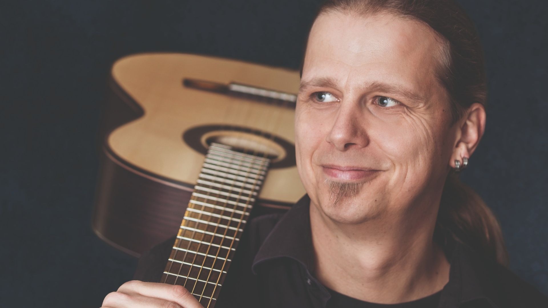 Lächelnder Mann mit Zopf und Goatee hält eine Akustikgitarre, die er mit der linken Hand am Hals greift