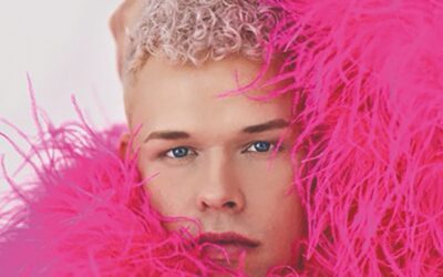 Tobias Born mit hellblauen Augen und kurzen, blonden Locken, umrahmt von leuchtend pinken Federn