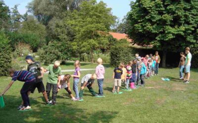 Kinder bewegen sich reihenweise auf grünen Matten über eine Wiese, umgeben von Bäumen und Sträuchern