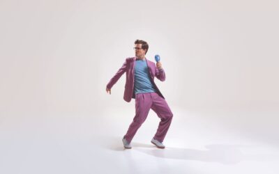 Bodo Wartke in lila Anzug und blauem Shirt hält blauen Handventilator in dynamischer Pose auf hellem Grund