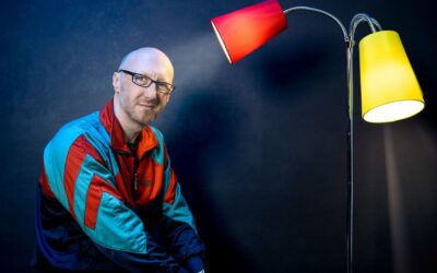Ralf Senkel mit Glatze und Brille im bunten Trainingsanzug lächelt vor dunklem Hintergrund neben einer Stehlampe