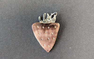 Erdbeerförmiger Metallanhänger mit kleinen Vertiefungen und silbernen Blättern auf dunklem Untergrund