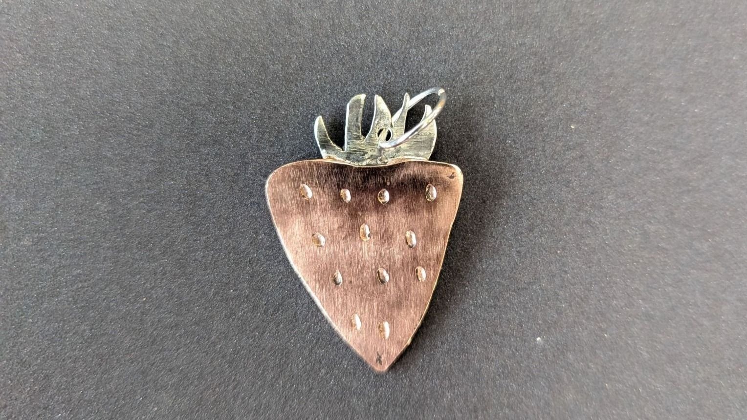 Erdbeerförmiger Metallanhänger mit kleinen Vertiefungen und silbernen Blättern auf dunklem Untergrund