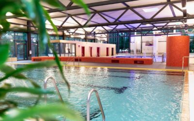 Großes, helles Hallenbad mit klarem Wasser, Edelstahlleitern, roten Kabinen und einer Sprunganlage im Hintergrund