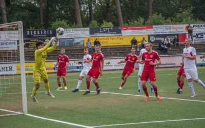 Ein Fußballtorwart in gelbem Trikot springt nach dem Ball vor dem Tor, umgeben von Spielern in roten und weißen Trikots