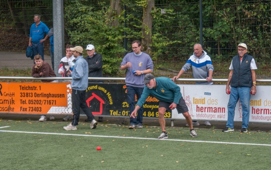 Männer spielen Boccia auf einem Sportplatz, ein Spieler bückt sich, um eine rote Kugel zu werfen, andere Spieler