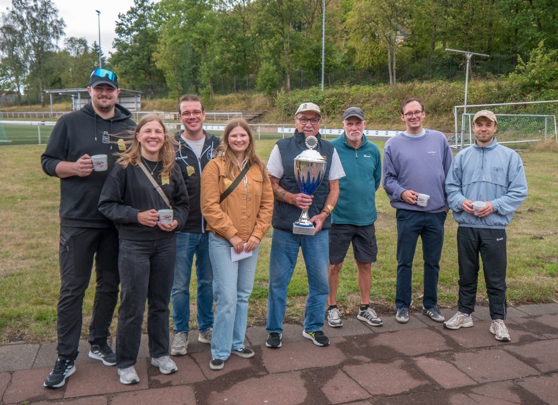 Sieben Personen des Teams Stukenbrock stehen lächelnd mit Tassen und dem Boccia-Pokal auf einem Sportplatz