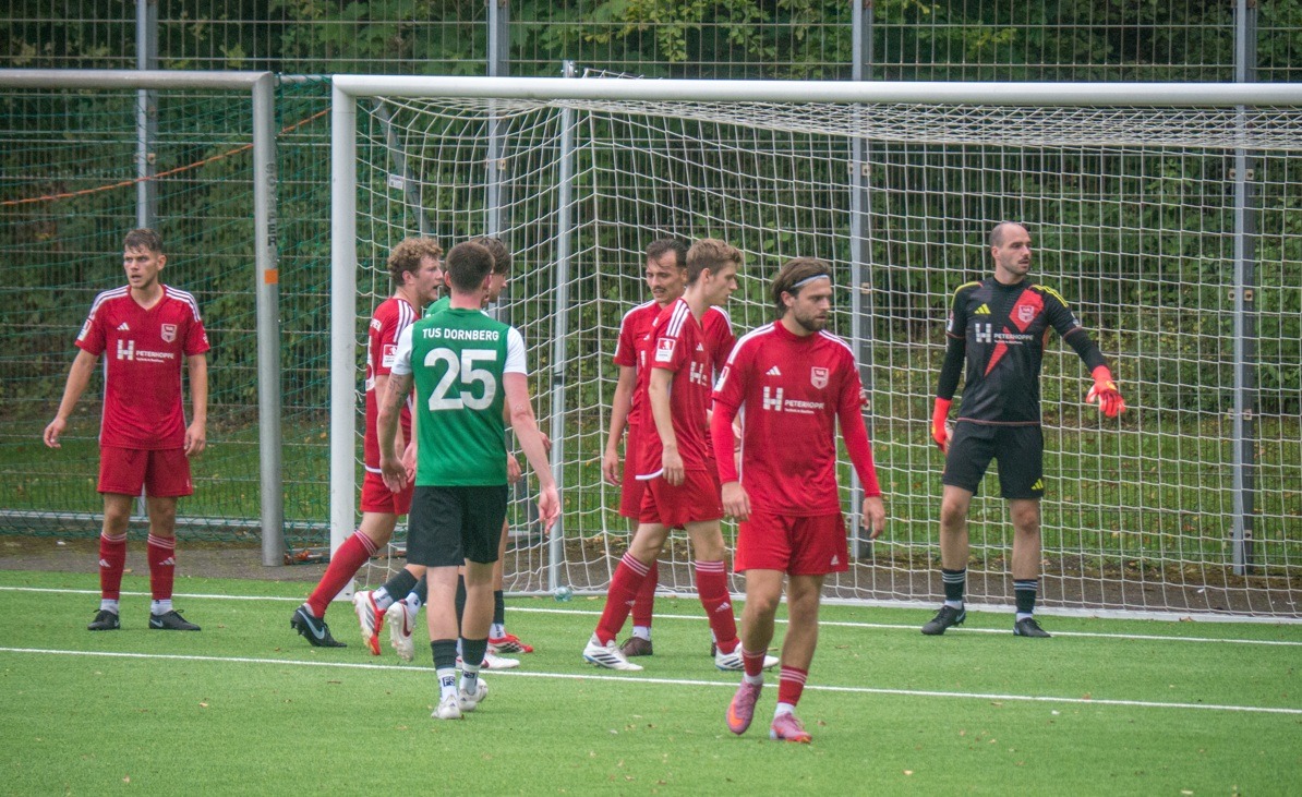 Mehrere Fussballspieler, die meisten in Rot, einer in Grün mit Dornberg 25, stehen auf einem grünen Feld vor einem Tor