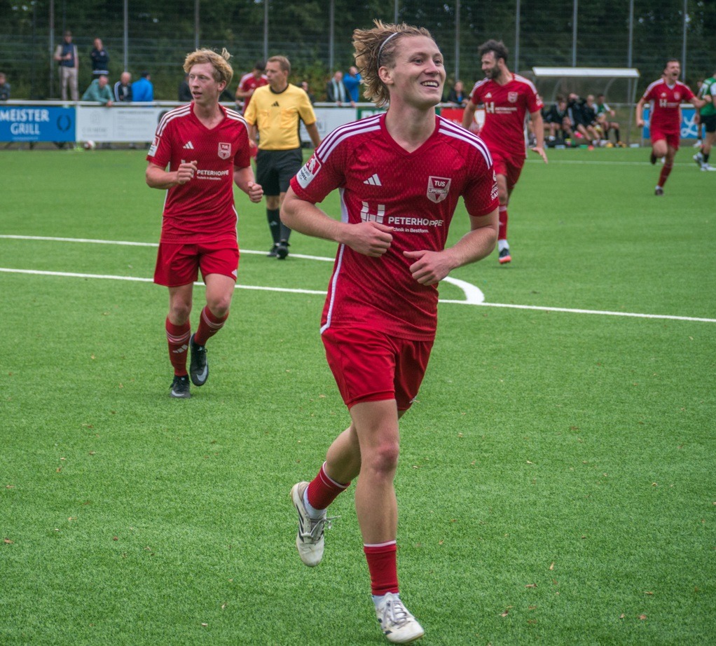 Lachender junger Fußballspieler in rotem Trikot des TuS Lipperreihe läuft auf grünem Kunstrasen, weitere Spieler folgen