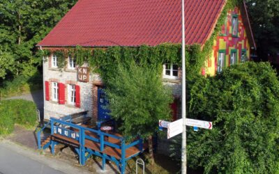 Zweigeteiltes Haus mit rotem Dach, eine Seite aus grauem Stein mit Efeu, die andere bunt bemalt, davor blaue Terrasse