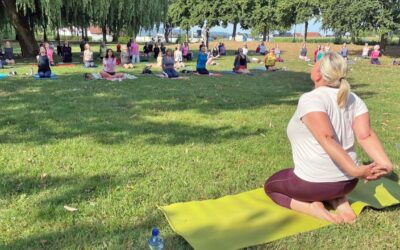 Zahlreiche Frauen machen Yoga auf Matten in einem sonnigen Park, kniend mit Händen hinterm Rücken und Blick nach oben