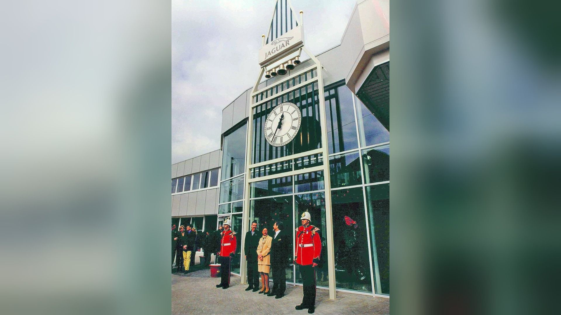 Eine große Big-Ben-artige Uhrfassade mit Jaguar-Logo vor einem modernen Gebäude, davor zwei Gardisten und drei Personen