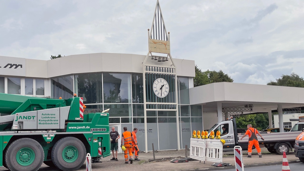 Mehrere Bauarbeiter und ein großer grüner Kran arbeiten an der Demontage eines Uhrturms mit Glocken vor einem Autohaus
