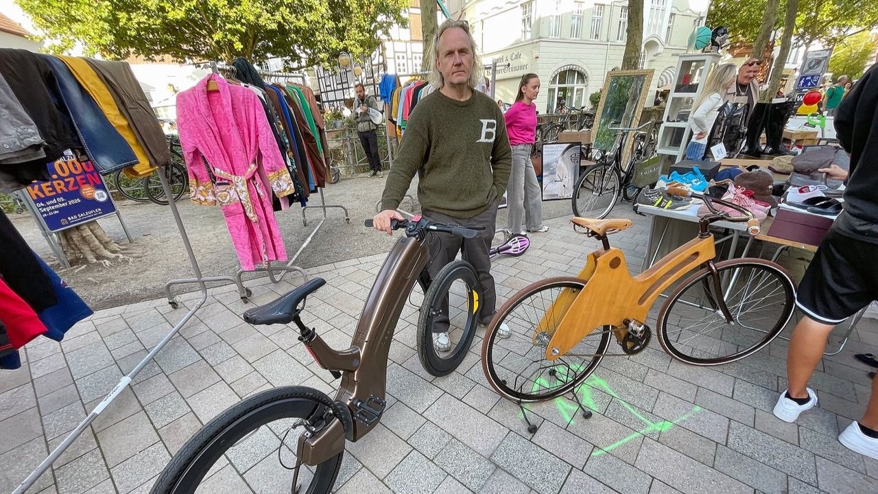 Mann steht neben einem braunen E-Bike und einem Holzfahrrad auf einem Außenmarkt mit Kleidern und Trödel