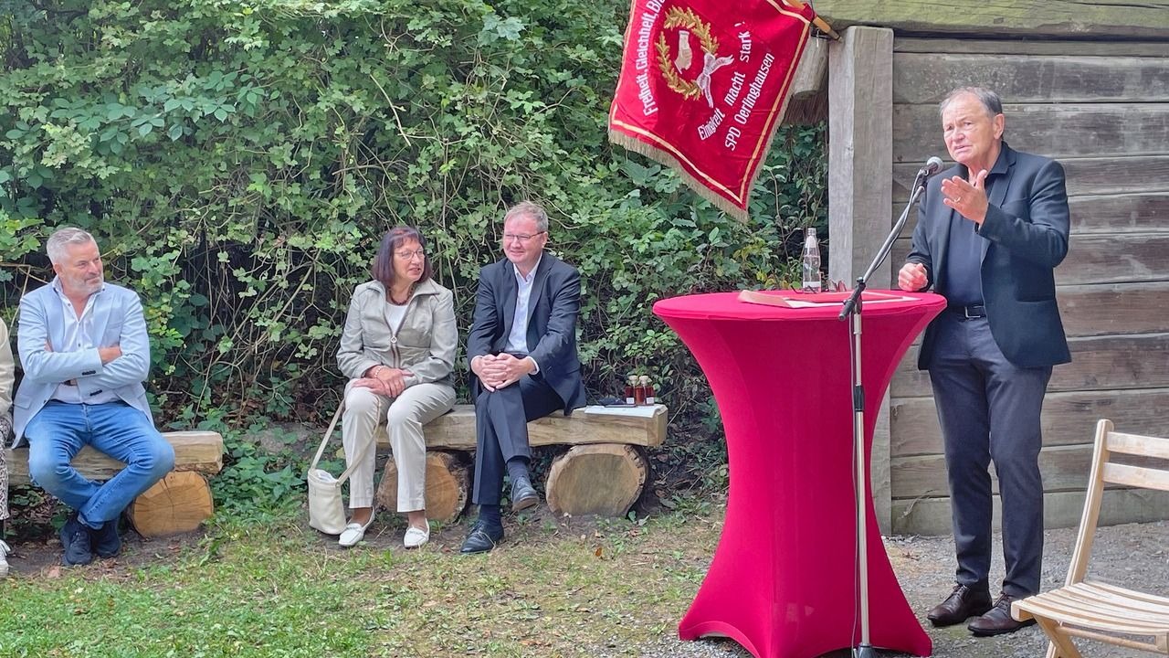 Ein Mann spricht an einem roten Stehtisch mit Mikrofon vor drei sitzenden Zuhörern und einem roten Banner der SPD