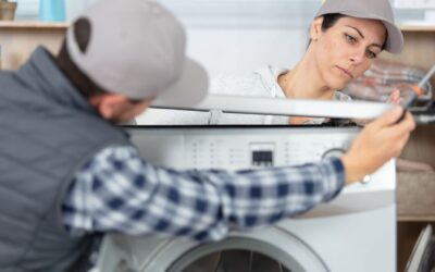 Zwei Personen mit Kappen reparieren gemeinsam eine Waschmaschine, die Frau hält einen Schraubenzieher