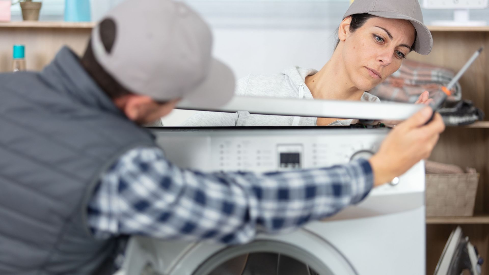 Zwei Personen mit Kappen reparieren gemeinsam eine Waschmaschine, die Frau hält einen Schraubenzieher