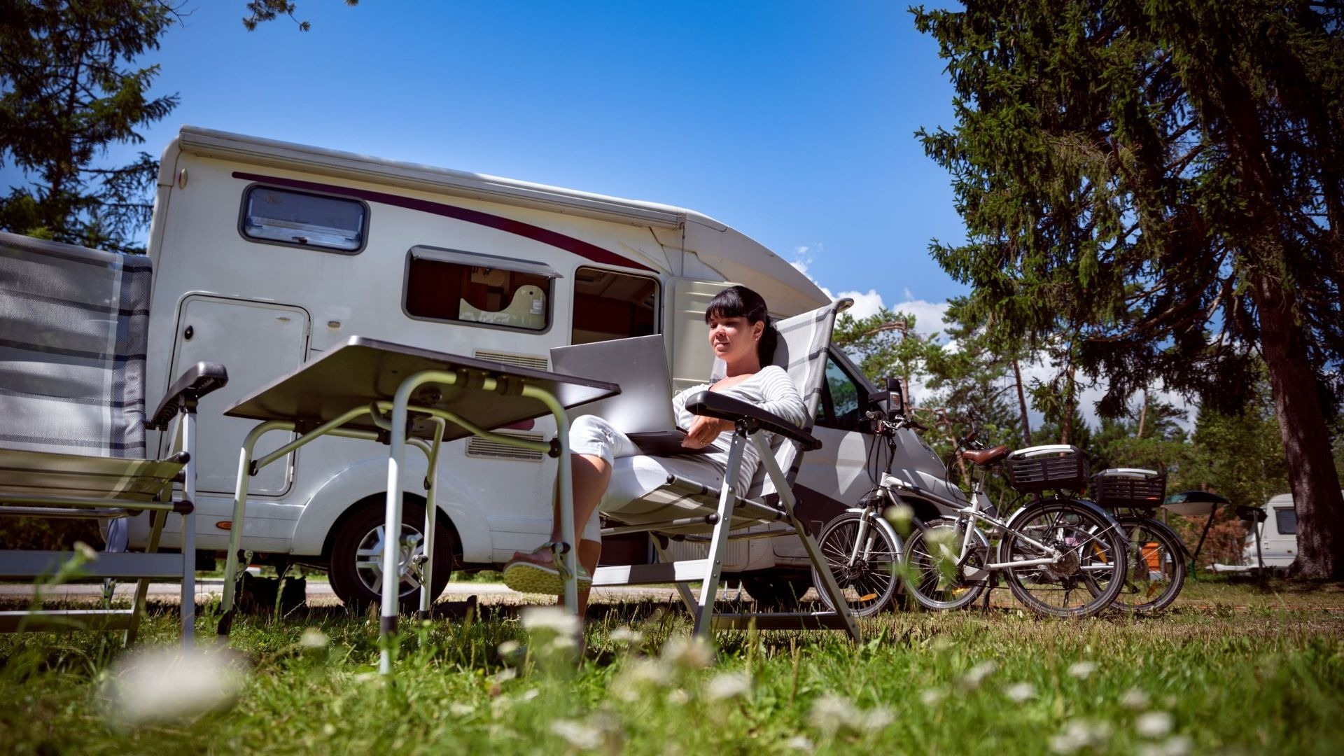 Frau sitzt mit Laptop auf einem Campingstuhl vor einem weißen Wohnmobil auf einer Wiese, daneben Fahrräder und Bäume
