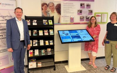 Ein Mann und zwei Frauen stehen lächelnd vor einem Infostand und einem digitalen Terminal zu Selbsthilfegruppen im