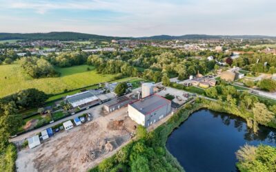 Luftaufnahme eines Industriekomplexes mit großen Gebäuden und Silos an einem dunklen Gewässer, umgeben von grüner