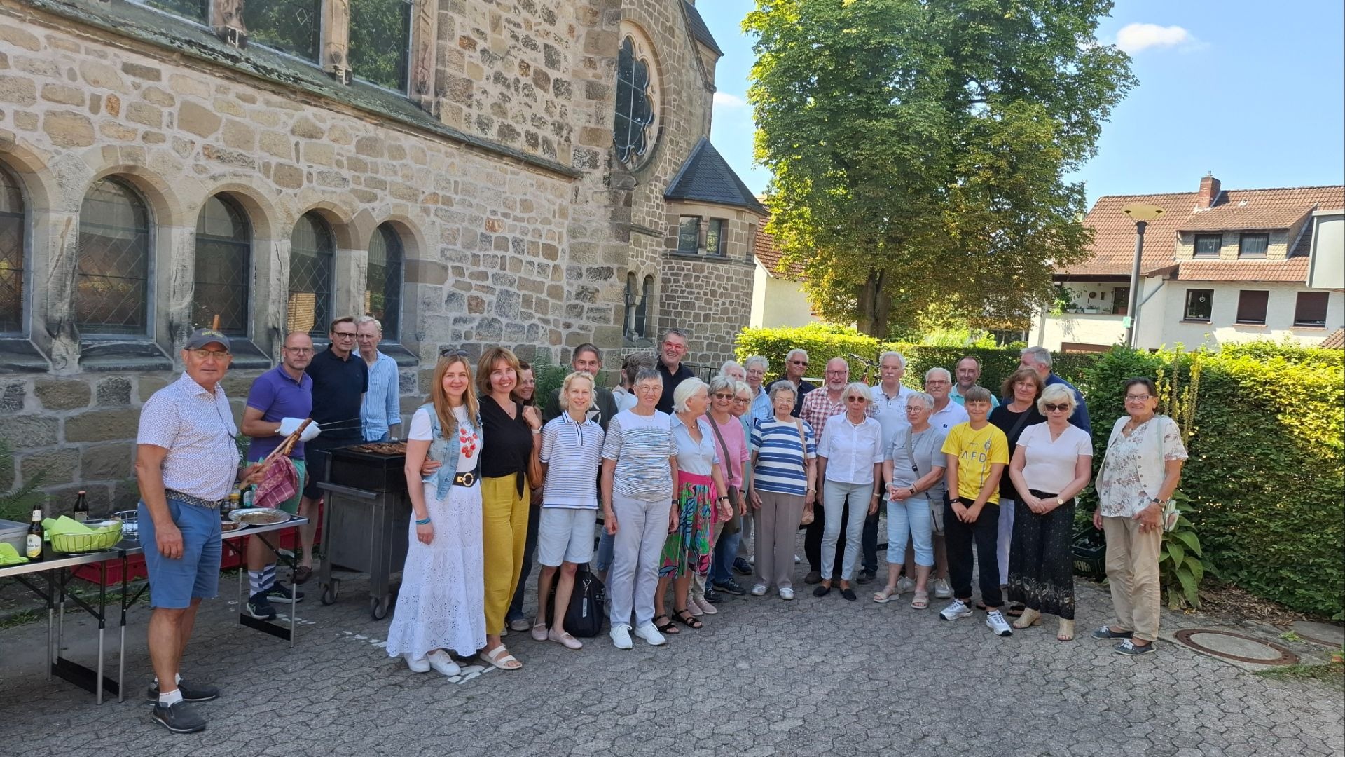 Viele Menschen stehen lächelnd vor einem Steingebäude, ein Mann grillt Würstchen an einem Grill neben Tischen mit