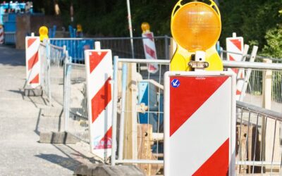 Baustelle mit rot-weißem Warnschild und gelbem Blinklicht, flankiert von Bauzäunen und weiteren Baken