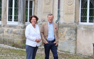 Kerstin Vieregge und ein Mann stehen lächelnd vor einem historischen Steingebäude auf gepflastertem Hof