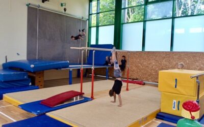 Turnhalle mit Kindern beim Training: Einer macht einen Handstand, einer schwingt hoch, andere sitzen auf Matten