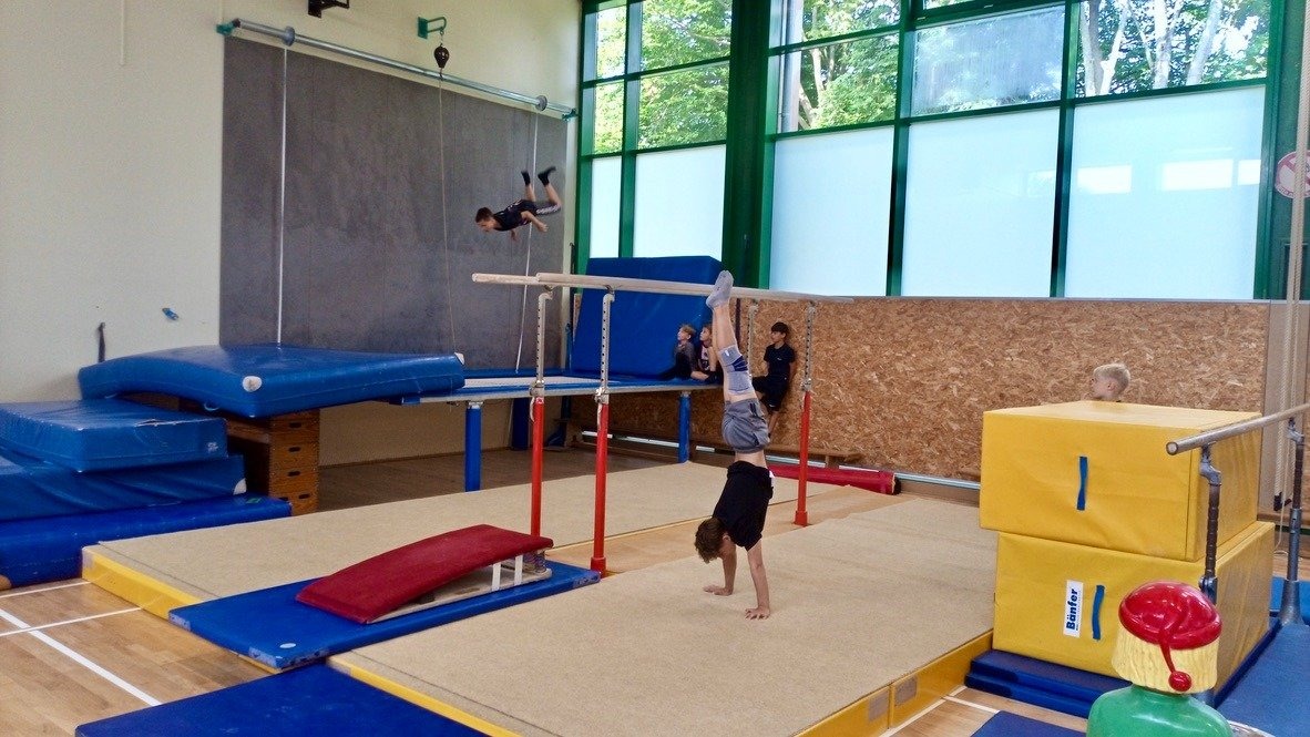 Turnhalle mit Kindern beim Training: Einer macht einen Handstand, einer schwingt hoch, andere sitzen auf Matten
