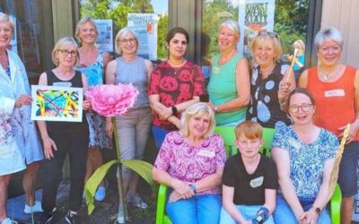 Gruppe Frauen und ein Junge posieren lächelnd draußen vor einem Gebäude, einige sitzen auf grüner Bank und zeigen