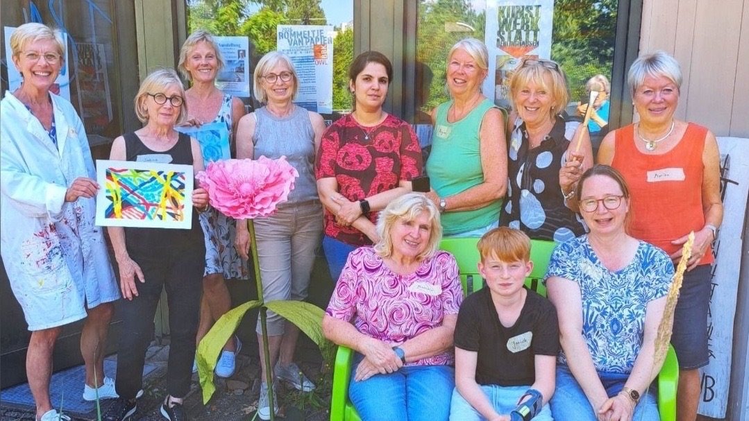 Gruppe Frauen und ein Junge posieren lächelnd draußen vor einem Gebäude, einige sitzen auf grüner Bank und zeigen