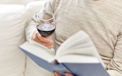 Eine Person hält ein Glas Rotwein und ein geöffnetes blaues Buch, sitzend auf einem weißen Sofa