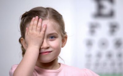 Kind bei Sehtest, bedeckt ein Auge mit der Hand, im Hintergrund eine unscharfe Sehtafel