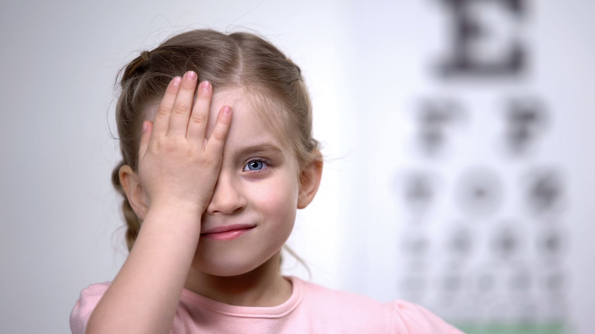 Kind bei Sehtest, bedeckt ein Auge mit der Hand, im Hintergrund eine unscharfe Sehtafel