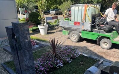 Friedhofsgärtner fährt auf grünem Bewässerungsfahrzeug an einem Grab mit Blumen und Grabstein vorbei