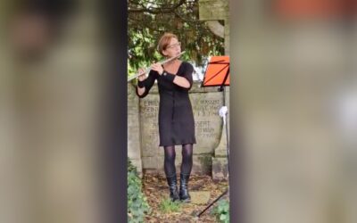 Frau in schwarzem Kleid spielt Querflöte vor einer beschrifteten Steinwand mit Notenständer