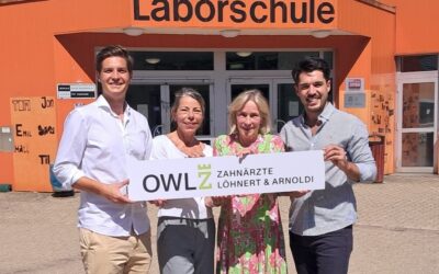 Vier lächelnde Personen halten ein Schild mit "OWL Z Zahnärzte Löhnert & Arnoldi" vor einem orangefarbenen Gebäude
