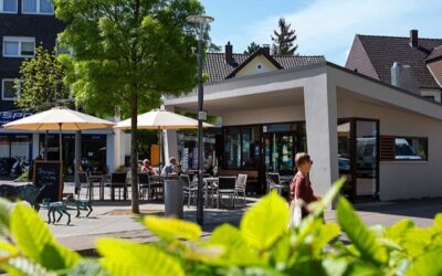 Modernes Café mit Gästen unter Sonnenschirmen auf einem Platz, davor eine Ziegenskulptur und eine Person geht vorbei