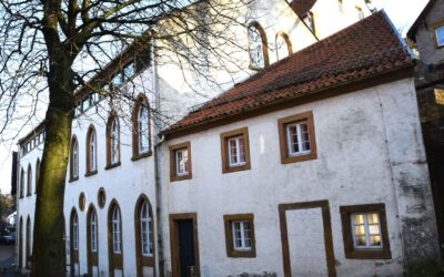 Alter, kahler Baum vor zwei historischen weißen Gebäuden mit roten Ziegeldächern und Rundbogenfenstern in