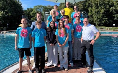 Gruppe von 16 Personen, viele in blauen T-Shirts mit Oktopus-Logo, stehen lächelnd am Schwimmbecken