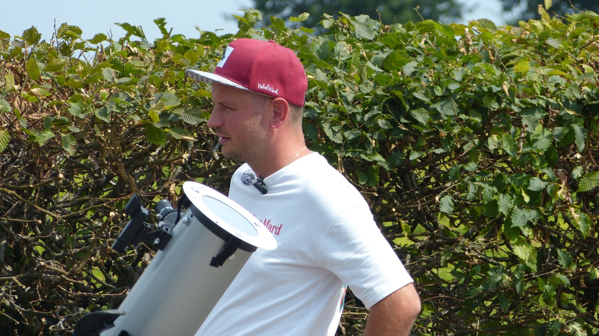 Mann mit roter Kappe trägt ein graues Teleskop mit Sonnenfilter vor einer grünen Hecke