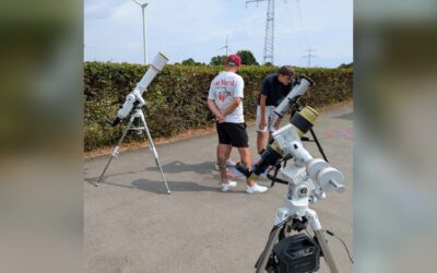 Zwei Männer mit drei aufgestellten Teleskopen auf einem Weg, einer schaut durch ein Teleskop, im Hintergrund Windräder