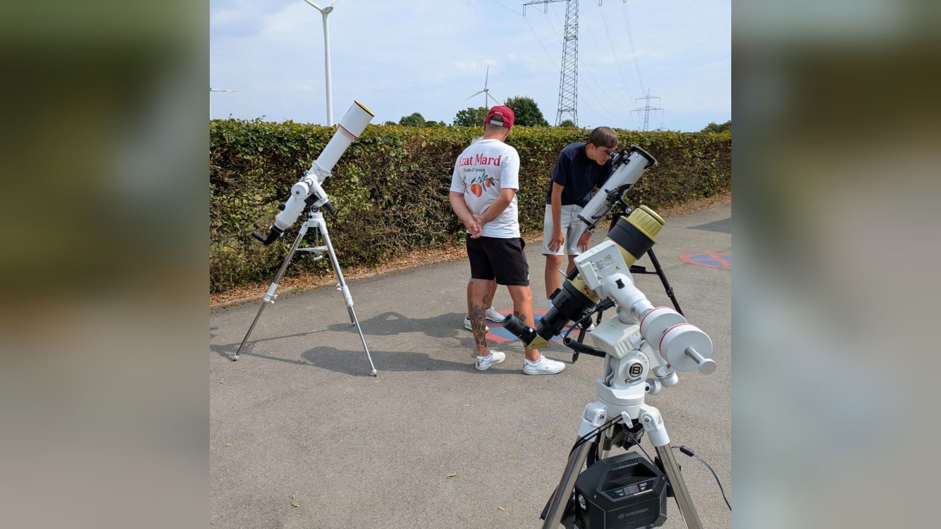 Zwei Männer mit drei aufgestellten Teleskopen auf einem Weg, einer schaut durch ein Teleskop, im Hintergrund Windräder