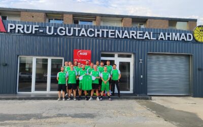 Gruppe Männer in grünen Trikots hält ein Trikot vor dem Gebäude „PRÜF- U. GUTACHTENAREAL AHMAD“