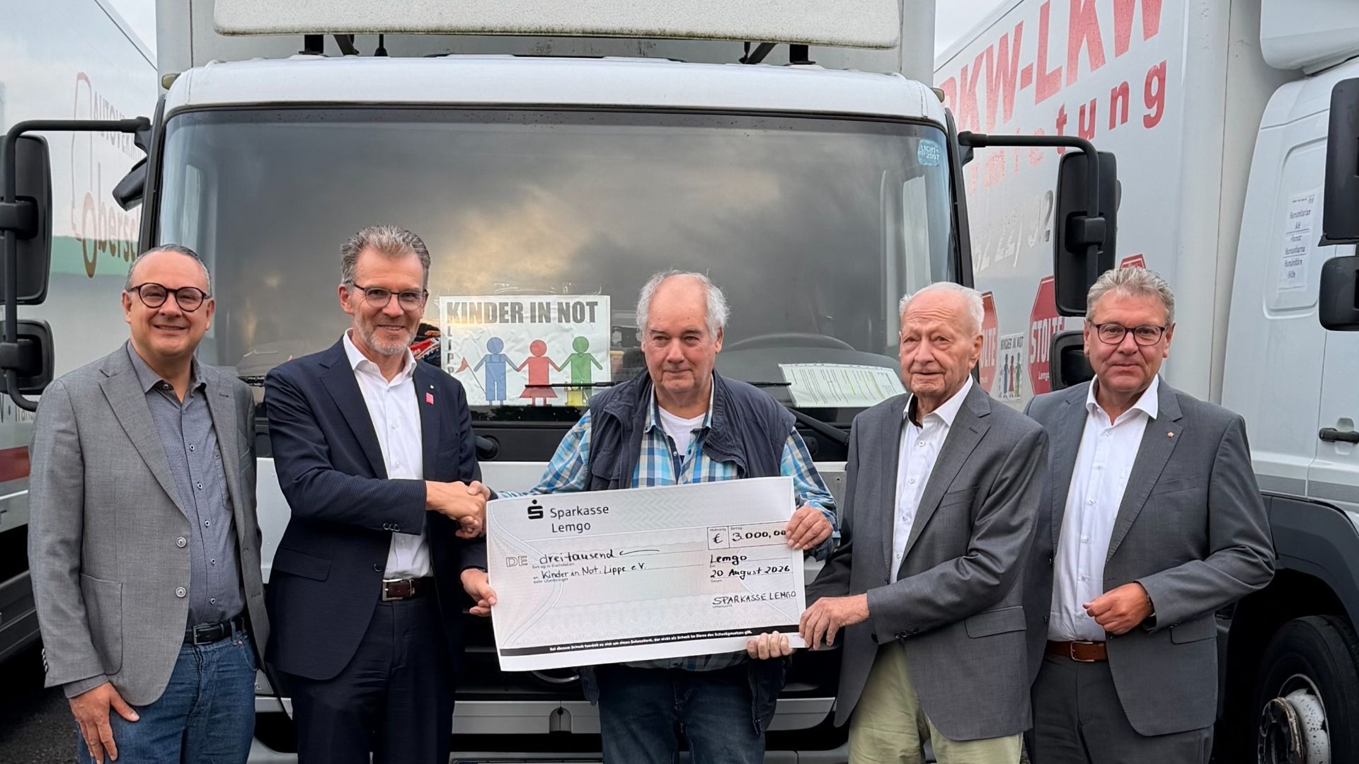 Fünf Männer halten einen Spendenscheck der Sparkasse Lemgo über 3.000 Euro vor Lastwagen der Organisation Kinder in Not