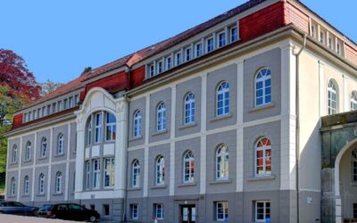 Gebäude in Oerlinghausen mit rotem Dach, grauer Fassade, Bogenfenstern und einem Erker, davor parken Autos