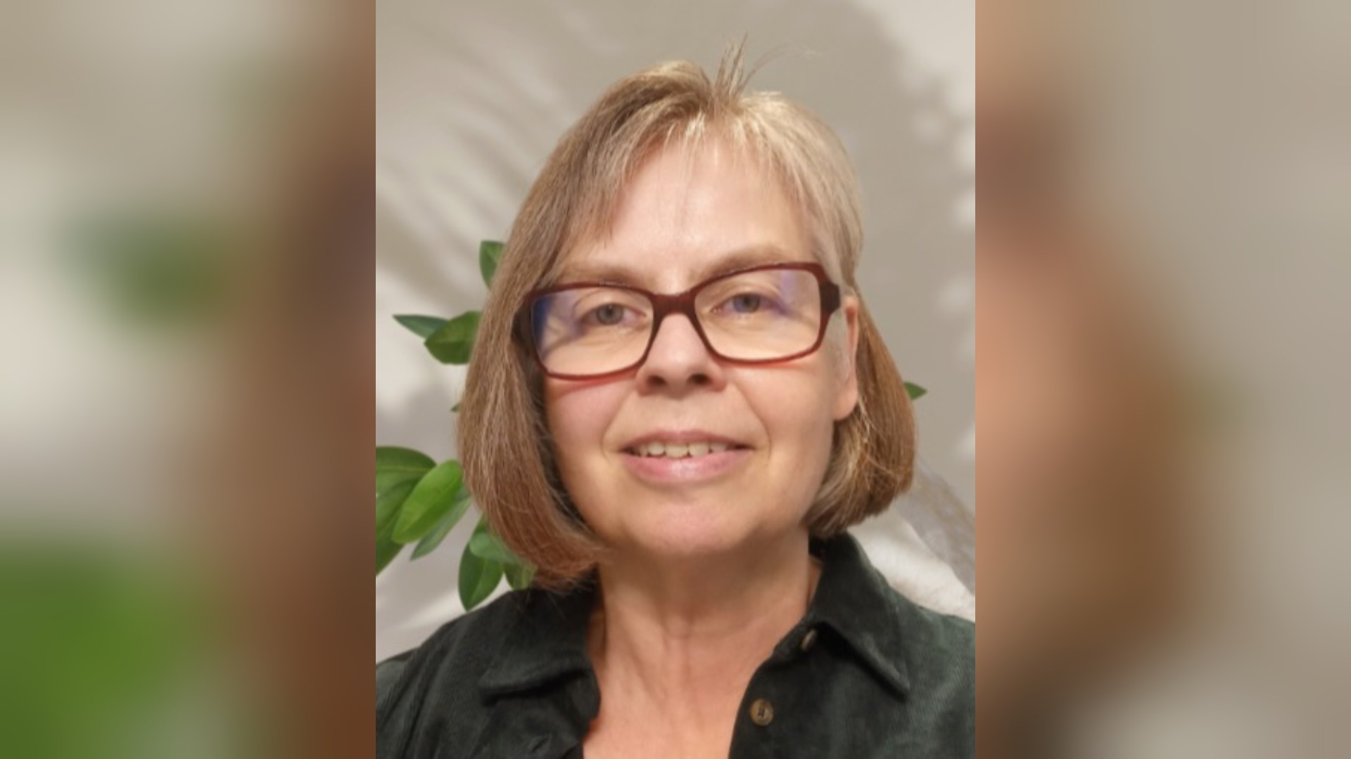 Eine lächelnde Frau mit kurzen braunen Haaren und roter Brille vor einer hellen Wand mit grünen Pflanzen