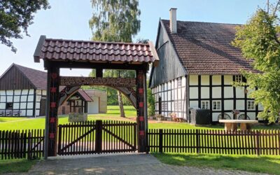 Großes Holztor mit Inschrift vor mehreren traditionellen Fachwerkhäusern und grüner Wiese unter blauem Himmel