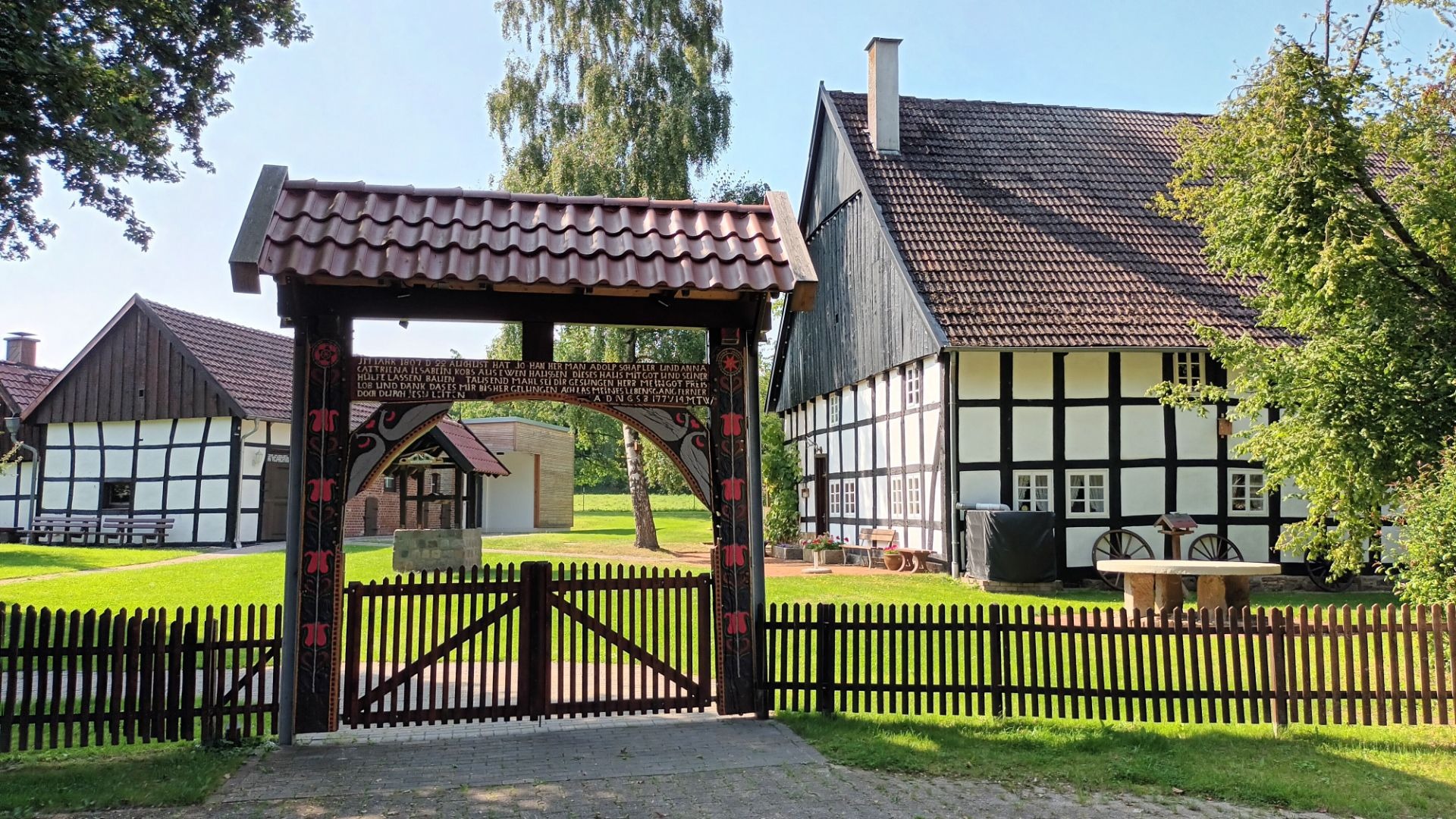 Großes Holztor mit Inschrift vor mehreren traditionellen Fachwerkhäusern und grüner Wiese unter blauem Himmel